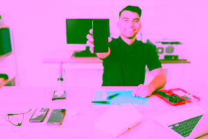 Eine Person sitzt an einem Schreibtisch in einer Reparaturwerkstatt und hält ein Smartphone hoch. Auf dem Schreibtisch befinden sich Werkzeuge, Telefonteile und ein Computer im Hintergrund.
