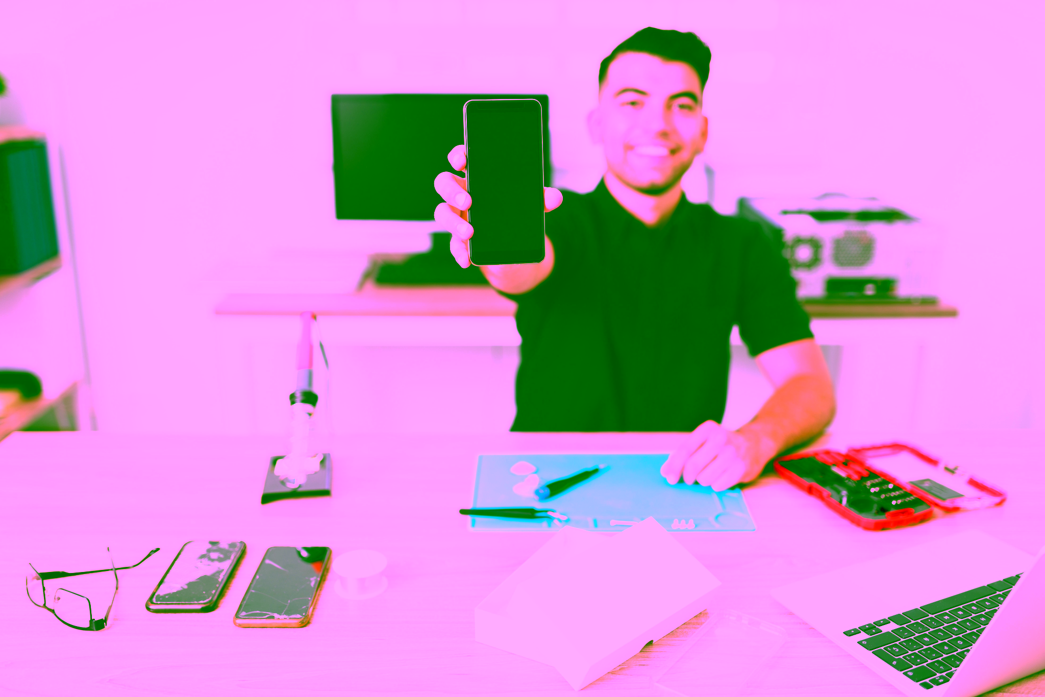 Eine Person sitzt an einem Schreibtisch in einer Reparaturwerkstatt und hält ein Smartphone hoch. Auf dem Schreibtisch befinden sich Werkzeuge, Telefonteile und ein Computer im Hintergrund.