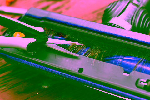 Nahaufnahme einer Schere, die Haare abschneidet, die sich um die Bürstenwalze eines Staubsaugers auf einer hölzernen Oberfläche verwickelt haben, und die eine laufende Reparatur eines Akkusaugers zeigt.