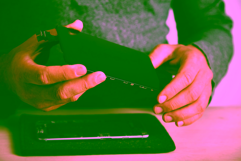 Eine Person hält einen abgetrennten Smartphone-Bildschirm über ein zerlegtes Telefon, das auf einer Matte liegt, und führt gekonnt eine Displayreparatur oder einen Bildschirmwechsel durch.