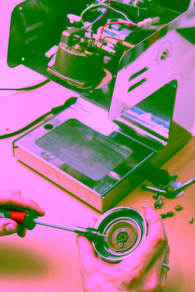 Person, die einen Schraubendreher benutzt, um den Siebträger einer Espressomaschine zu reparieren oder zu demontieren, wobei im Hintergrund interne Maschinenteile sichtbar sind.